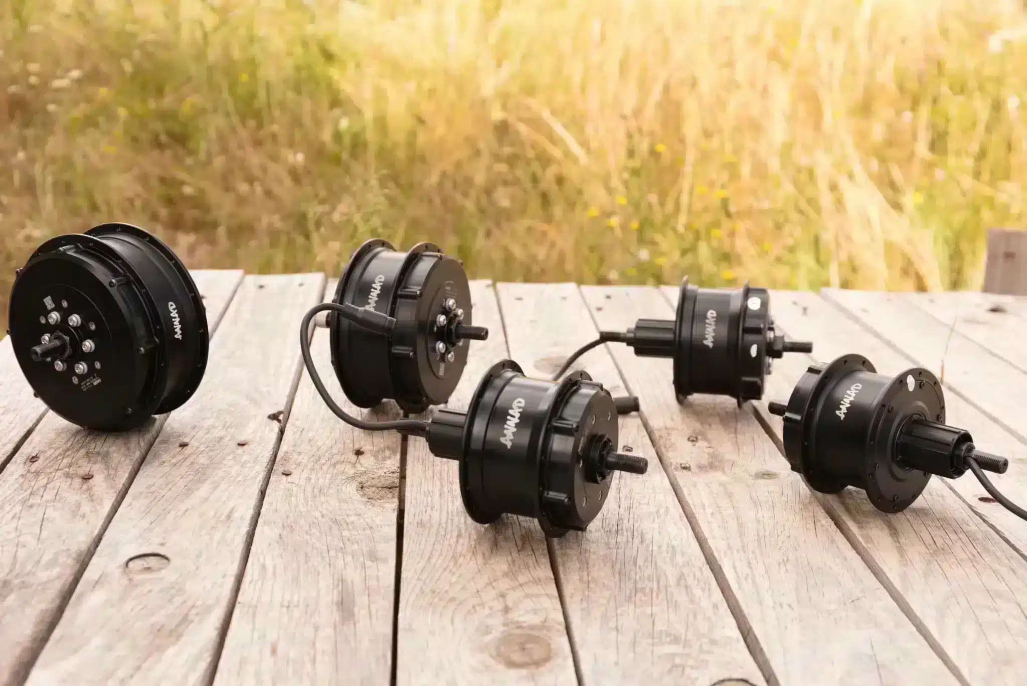Moteur roue avant , roue arrière et pédalier : comparons-les ! 1 Cinq moteurs de roue de vélo ANNAD sur une palette en bois avec une base en herbe. De gauche à droite : deux moteurs ALL IN ONE avec batterie intégrée, un moteur CLASSIC et deux moteurs ULTRA.